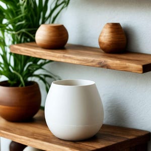 Puede incluir: Estanterías flotantes de madera con varios objetos decorativos. Las estanterías exhiben pequeñas plantas en macetas, jarrones de madera y un jarrón blanco grande. Las estanterías son de madera oscura y están montadas en una pared blanca.
