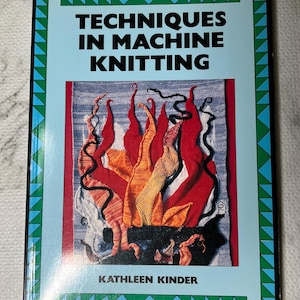 May include: A book titled "Techniques in Machine Knitting" by Kathleen Kinder. The cover features a colorful textile art piece with red, orange, and yellow flame-like shapes against a blue background. The book has a light blue border with green and black triangles.