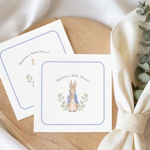 May include: Two square baby shower invitations with a blue border and the text "Madeline's Baby Shower." One invitation features a watercolor illustration of Peter Rabbit holding a carrot. The invitations are on a wooden tray.