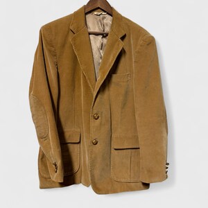 May include: A tan corduroy blazer with a notched lapel, two front pockets, and elbow patches. The blazer has a two-button closure and is hanging on a wooden hanger. The lining is visible inside the blazer.