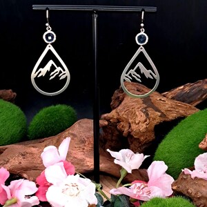 May include: A pair of silver-toned drop earrings featuring a teardrop-shaped frame with a cutout mountain range design. Each earring is accented with a small, round, dark blue faceted stone at the top, suspended from a silver-toned hook wire.