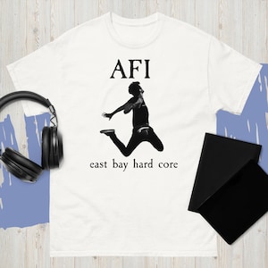 May include: White t-shirt featuring the band name "AFI" in black capital letters above a black and white image of a person jumping. The text "east bay hard core" is printed below the image in black.