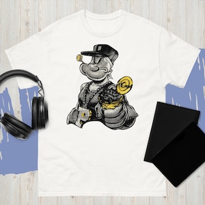 Peut inclure: T-shirt blanc avec une illustration en noir et blanc de Popeye le marin, portant une chaîne en or, un chapeau et une sucette. Le personnage de dessin animé tient un récipient doré. Le t-shirt a un col rond et des manches courtes.