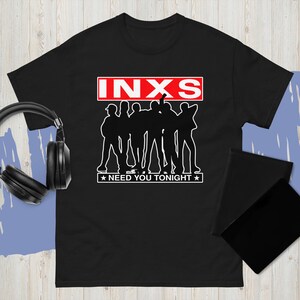 May include: Black t-shirt featuring the band INXS. The design includes the band's name in a red box, a silhouette of the band members, and the text "NEED YOU TONIGHT" with stars. A pair of headphones and a black notebook are visible.