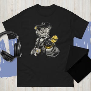 Peut inclure: T-shirt noir avec une illustration de Popeye le marin dans un style gangster. Le graphisme est en nuances de gris et de jaune, avec Popeye portant une casquette et tenant une sucette. Un design original.