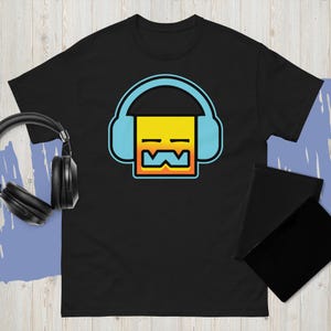 May include: Black t-shirt featuring a geometric design of a yellow square with blue headphones and a stylized mouth. The design has orange and blue accents. A pair of black headphones is visible in the background.