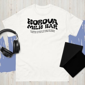 May include: White t-shirt with the text "KOROVA MILK BAR" in a bold, black font. Below the main text is the phrase "SHARPEN YOURSELF UP AND GET READY FOR A BIT OF THE OLD ULTRA-VIOLENCE" in a smaller, black font. A pair of black headphones and two black notebooks are also visible.