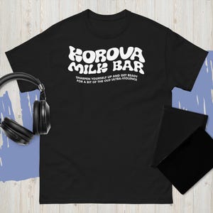 May include: Black t-shirt with the text "KOROVA MILK BAR" in white, along with the phrase "SHARPEN YOURSELF UP AND GET READY FOR A BIT OF THE OLD ULTRA-VIOLENCE". The shirt is short-sleeved and has a crew neck.