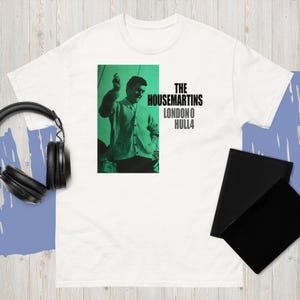 以下が含まれることがあります： カーディガンを着た男性の緑と黒のグラフィックが特徴の白いTシャツ。グラフィックには「THE HOUSEMARTINS LONDON O HULL4」というテキストが含まれています。黒いヘッドフォンと黒いノートも表示されます。