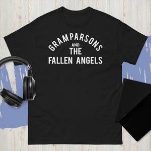 Könnte beinhalten: Schwarzes T-Shirt mit dem weißen Schriftzug "Gram Parsons and The Fallen Angels" in Bogenform. Das T-Shirt wird auf einer hellen Holzoberfläche präsentiert, mit schwarzen Kopfhörern und einem schwarzen Tablet im Hintergrund.