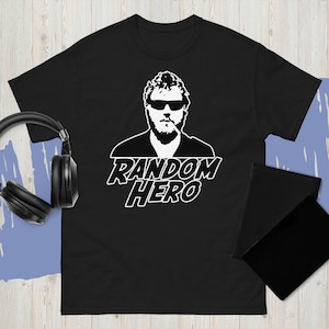 Può includere: T-shirt nera con grafica bianca di una persona con occhiali da sole e barba, con la scritta "RANDOM HERO" sotto. Sono visibili delle cuffie nere e un blocco note nero.