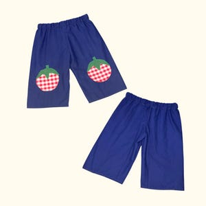 May include: Two pairs of blue, wide-leg trousers. One pair features a red and white gingham apple appliqué on each knee, with green leaf details. The other pair is plain blue. Both have elasticated waistbands.