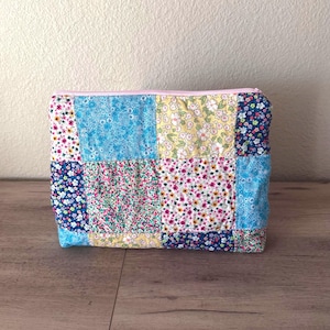 Puede incluir: Un neceser rectangular de patchwork con cremallera rosa claro. La tela tiene estampados de pequeñas flores en azul, rosa y amarillo. El neceser parece estar acolchado.