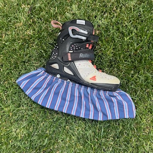 Peut inclure: Un patin à roulettes noir et gris avec des touches corail et le mot "Rollerblade". Le patin est posé sur une housse en tissu rayé bleu et blanc, sur de l'herbe verte.