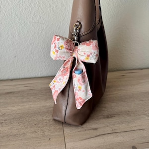 May include: A floral bow keychain with pink and yellow flowers is attached to a brown leather handbag. The bow has long, flowing ribbons and a small pocket holding a blue item. The bag is on a wooden surface.