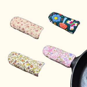 May include: Four fabric pan handle covers in various floral patterns. One is cream with pink flowers, another is dark green with a butterfly and flowers, a third is yellow with orange and brown flowers, and the last is pink with purple flowers.