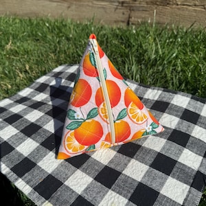 May include: A triangular zippered pouch with a vibrant orange fruit pattern on a pink background. The pouch is quilted and features a beige zipper. It rests on a black and white checkered cloth, with green grass in the background.