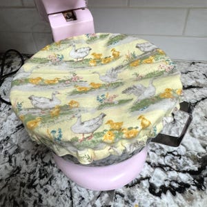 Op de afbeelding: Een lichtgele stoffen hoes met een eenden- en eendenkuikenpatroon, die een roze keukenmixer bedekt. De hoes heeft illustraties van witte ganzen, gele eendjes en bloemaccenten. De mixer staat op een aanrecht.