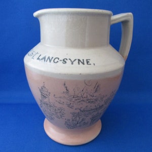 Peut inclure: Pichet en céramique vintage, rose clair et blanc. Il présente une illustration de paysage bleue et le texte "St Lanc-Syne". Il est doté d'une anse et d'un socle, typique des objets anciens.