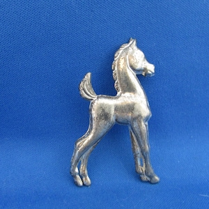 Könnte beinhalten: Eine silberfarbene Metallbrosche in Form eines stehenden Fohlens. Das Fohlen hat eine detaillierte Mähne und einen Schweif, und seine Beine sind schlank. Die Brosche ist vor einem einfarbigen blauen Hintergrund platziert.