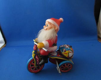 May include: A vintage toy featuring Santa Claus riding a tricycle. Santa is dressed in a red suit and hat with a white beard. The tricycle is red, blue, and yellow with a helmet on the back. A classic Christmas collectible.