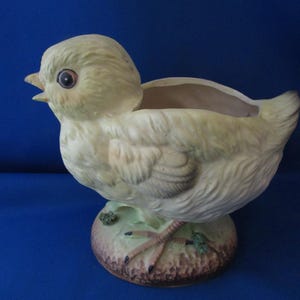 Puede incluir: Un macetero de cerámica con forma de pollito con un esmalte amarillo y blanco. El pollito tiene un pico marrón y ojos negros. El macetero tiene una base marrón con detalles verdes y marrones.