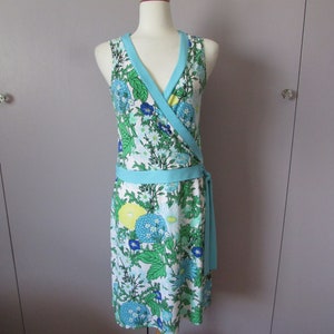 May include: A sleeveless wrap dress with a floral pattern in shades of blue, green, and yellow on a white background. The dress features a light blue trim and a matching waist tie. The dress is displayed on a mannequin.