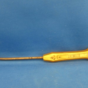 May include: A vintage gold-colored metal hook tool with a curved end and a decorative handle. The handle has floral designs and text. The tool is set against a blue background.