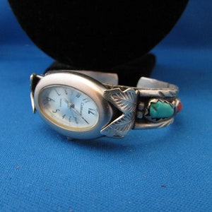 Op de afbeelding: Zilverkleurige horloge met een witte wijzerplaat en zwarte wijzers. De horlogeband is een zilveren armband met turquoise en rode stenen en een bladontwerp.