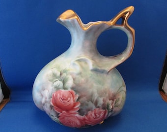 May include: A hand-painted ceramic jug with a bulbous body and a curved handle. The jug features a light blue and green background with pink roses and gold trim. The jug is approximately 20 cm tall.