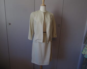 May include: A cream-coloured vintage skirt suit on a mannequin. The suit features a collared jacket with three-quarter sleeves and a matching knee-length skirt. The jacket has a subtle checkered pattern and button details.
