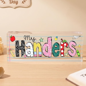 Könnte beinhalten: Ein rechteckiger Schreibtischaufsteller aus klarem Acryl mit dem Namen Mrs. Handers, der in bunten, gemusterten Buchstaben gedruckt ist. Das Design enthält schulbezogene Grafiken wie einen roten Apfel, einen gelben Stift und Sterne.
