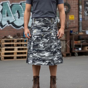 May include: A gray and white camouflage kilt with brass-colored buttons and pockets. The kilt is worn with a gray t-shirt and brown boots. The kilt is a traditional Scottish garment, often worn for formal occasions or as a statement piece.