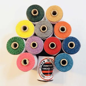 Pode incluir: Uma coleção de bobinas de linha de linho coloridas em vários tons, incluindo verde, amarelo, laranja e azul. Uma bobina cilíndrica de cordão Crawford Threads Ltd. também é visível. As bobinas estão dispostas em um padrão decorativo.
