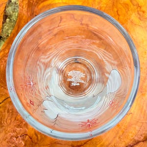 May include: Clear glass tumbler with a design featuring a white outline of a bird and red starburst accents. The bottom of the glass has text and a logo. The glass is empty and viewed from above.