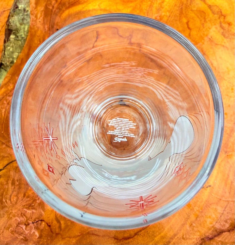 May include: Clear glass tumbler with a design featuring a white outline of a bird and red starburst accents. The bottom of the glass has text and a logo. The glass is empty and viewed from above.