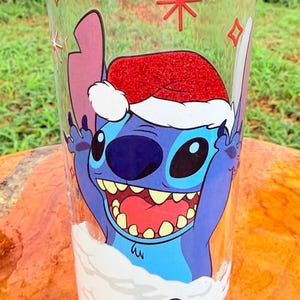May include: A clear glass featuring a cartoon character wearing a Santa hat. The character is blue with large eyes and a wide smile. The glass has a festive design with snowflakes and a red star.