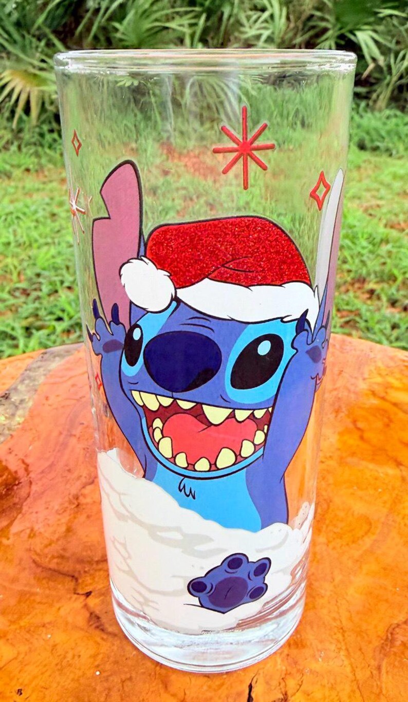 May include: A clear glass featuring a cartoon character wearing a Santa hat. The character is blue with large eyes and a wide smile. The glass has a festive design with snowflakes and a red star.