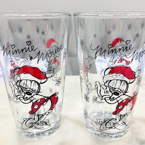 Pu&ograve; includere: Due bicchieri trasparenti con un design di Minnie Mouse. I bicchieri presentano un'illustrazione a cartoni animati di Minnie Mouse con un cappello da Babbo Natale rosso e una gonna a pois rossi. Le parole "Minnie Mouse" sono scritte in corsivo.