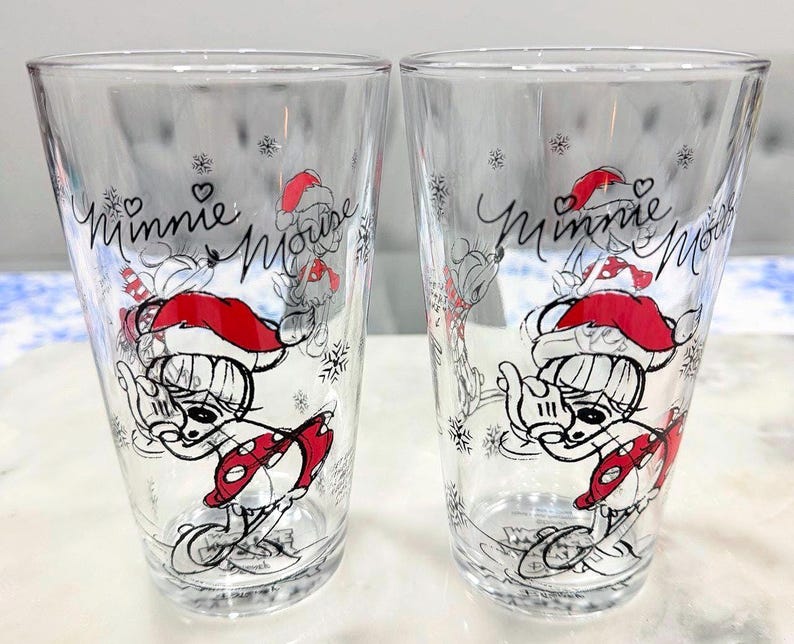 Pu&ograve; includere: Due bicchieri trasparenti con un design di Minnie Mouse. I bicchieri presentano un'illustrazione a cartoni animati di Minnie Mouse con un cappello da Babbo Natale rosso e una gonna a pois rossi. Le parole "Minnie Mouse" sono scritte in corsivo.