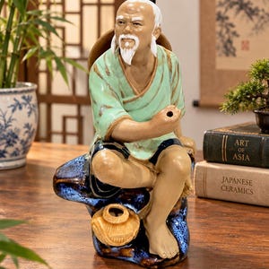 May include: A ceramic figurine of an elderly person with a white beard and topknot, wearing a light green tunic and dark shorts. The figure sits on a textured blue and brown rock base next to a small tan pot, with one hand raised near the face.