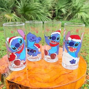 May include: Four clear drinking glasses with cartoon character Stitch designs. Each glass features a different Stitch pose, wearing a Santa hat and red scarf. The glasses are arranged on a wooden surface, with a green background.