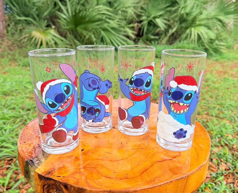 May include: Four clear drinking glasses with cartoon character Stitch designs. Each glass features a different Stitch pose, wearing a Santa hat and red scarf. The glasses are arranged on a wooden surface, with a green background.