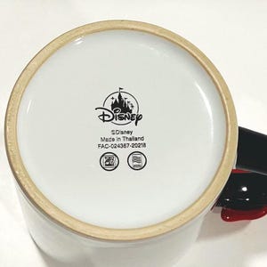 Puede incluir: Taza de cer&aacute;mica blanca con asa negra y detalles rojos. La base de la taza presenta el logotipo de Disney, el texto "Disney", "Made in Thailand" y otras marcas.