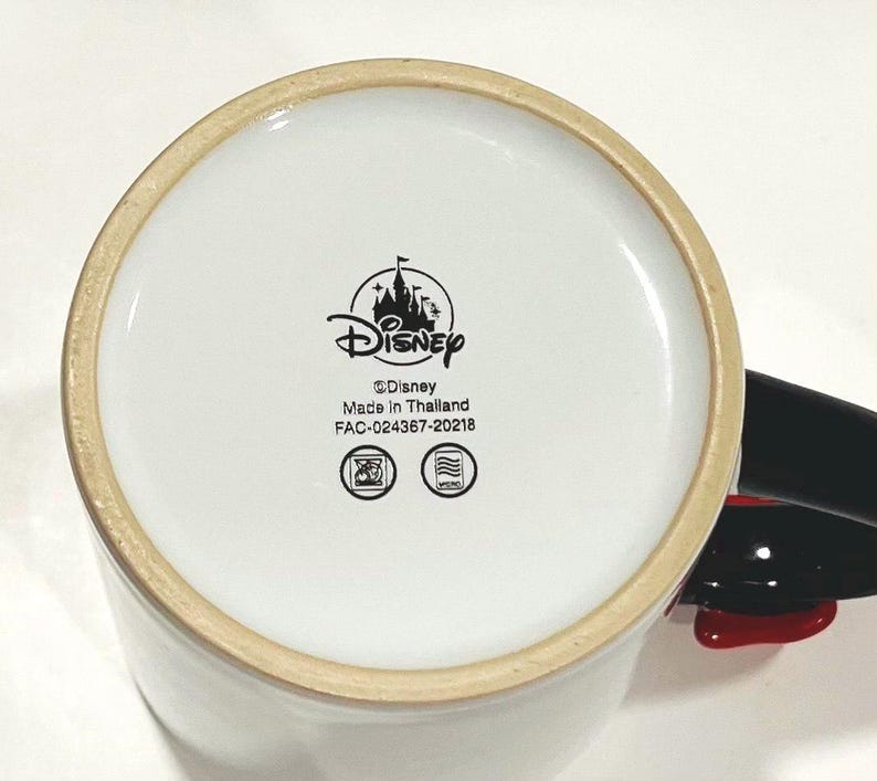 Puede incluir: Taza de cer&aacute;mica blanca con asa negra y detalles rojos. La base de la taza presenta el logotipo de Disney, el texto "Disney", "Made in Thailand" y otras marcas.