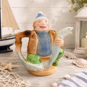 May include: A ceramic teapot in the shape of a fisherman holding a large fish. The teapot features a smiling fisherman wearing a blue hat, a blue and white striped sweater, and a brown jacket. The fish is light blue with detailed scales.