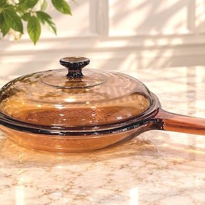 May include: A round, amber-coloured glass skillet with a matching transparent glass lid. The pan features a long, integrated glass handle and a circular knob on the centre of the lid. The glass has a smooth, translucent finish with a warm brown tint.