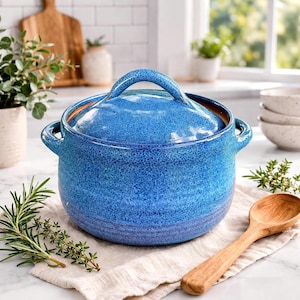 May include: A blue ceramic Dutch oven with a lid and side handles. The pot has a speckled texture and a glossy finish. A wooden spoon and sprigs of rosemary and thyme are nearby. The pot is sitting on a white cloth.