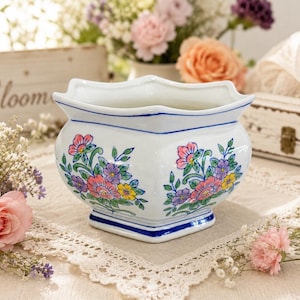 May include: A white ceramic planter with a scalloped rim and a square base, featuring hand-painted floral motifs in shades of pink, purple, and yellow with green leaves. The vessel is accented with thin blue lines along the rim and base edges.