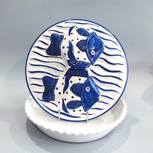 May include: A white ceramic plate with a blue fish design. The plate features two fish, wave patterns, and decorative elements. The plate is displayed on a white, scalloped-edge bowl. The design is in a nautical theme.